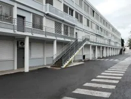 Immeuble moderne avec escaliers extérieurs, façades grises, fenêtres à persiennes et route d’accès bien marquée.