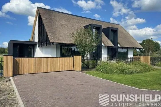 Maison moderne avec toit de chaume, façade blanche, éléments noirs et clôture en bois, entourée de verdure et ciel dégagé.