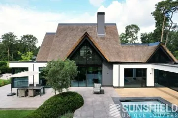 Maison moderne avec un toit en chaume, piscine et terrasse, entourée d’un jardin verdoyant et arboré.