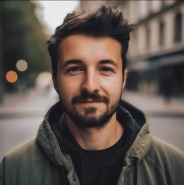 Portrait d’un jeune homme souriant, avec une barbe et des cheveux bruns, en veste, devant un décor urbain flou.