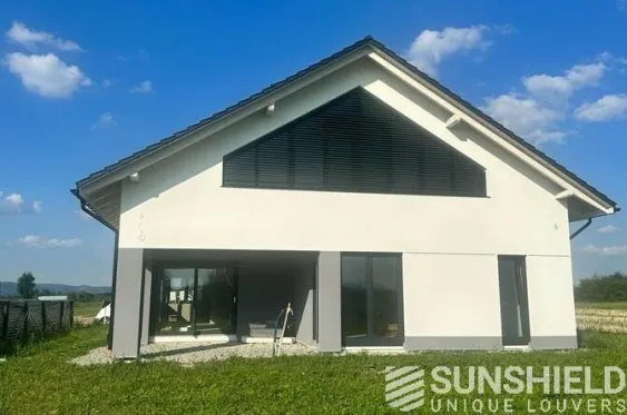Maison moderne à toit mansardé, fenêtres larges, située dans un environnement naturel verdoyant sous un ciel bleu.
