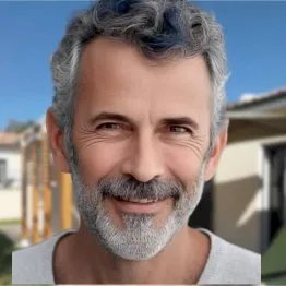 Homme souriant avec des cheveux gris et une barbe, devant un décor extérieur. Ambiance conviviale et détendue.