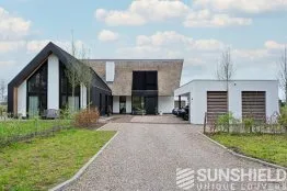 Maison moderne avec façade en bois et grandes fenêtres, garage attenant, entourée de verdure et d’une allée en gravier.