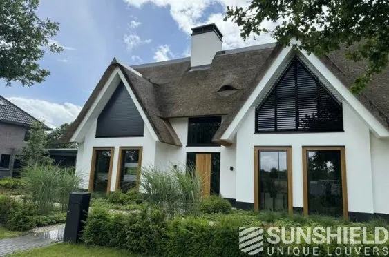 Maison moderne au toit de chaume, avec fenêtres en bois et volets, entourée de verdure et d’un jardin soigné.