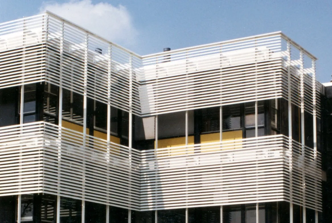 Façade moderne d’immeuble avec volets en lattes blanches et fenêtres aux accents jaunes, sous un ciel bleu clair.