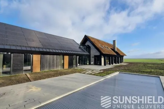 Maison moderne avec panneaux solaires, grandes baies vitrées et toiture en pente, située dans un paysage verdoyant.