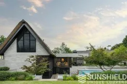 Maison moderne avec toiture en pente, façade en briques et piscine, entourée de verdure et d’arbres, baignées de lumière naturelle.