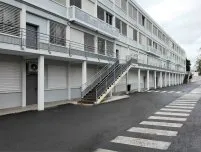 Immeuble moderne avec escaliers extérieurs, façades grises, fenêtres à persiennes et route d’accès bien marquée.