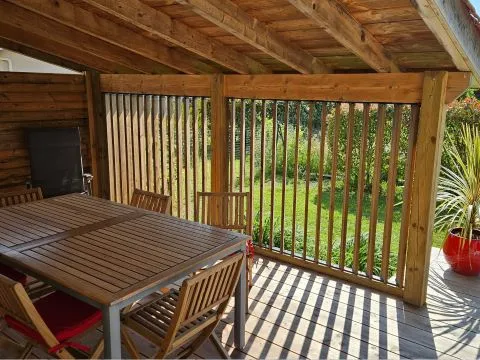 Terrasse en bois avec table, chaises et verdure, sous un abri avec des lames en bois, créant une ambiance chaleureuse.