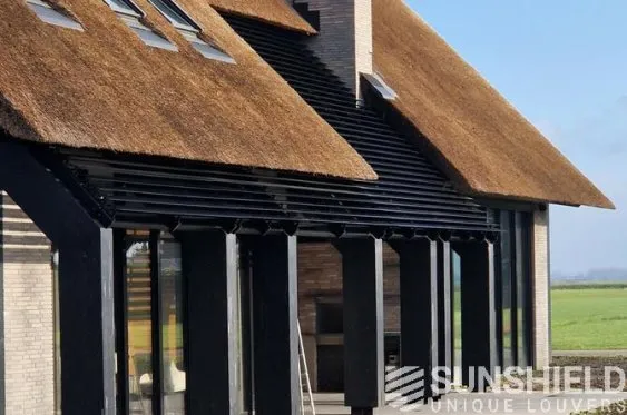 Maison moderne avec toiture en chaume, façades vitrées et lames d’ombre en bois, situé dans un paysage verdoyant.